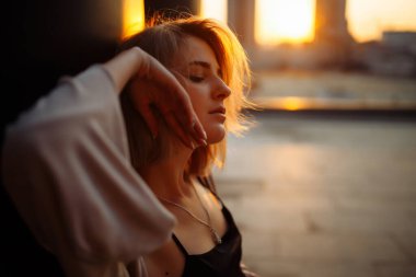 Çatıda fotoğraf çekimi. Gün batımında çatıda poz veren genç bir kadın. Moda, tarz kavramı.