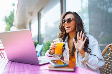 Terasta genç ve neşeli bir kadın Skype 'ta sohbet ediyor, webcam' de konuşuyor, video konferansı yapıyor. Kadın modern bir kafede oturuyor ve bir dizüstü bilgisayar üzerinde çalışıyor. Serbest Çalışan, Eğitim Konsepti.
