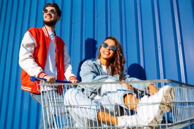 Güzel arkadaşlar açık havada alışveriş arabalarında eğleniyorlar. Yaşam tarzı, eğlence, eğlence, gençlik kavramı. Tarz sahibi erkek ve kadın eğleniyor..