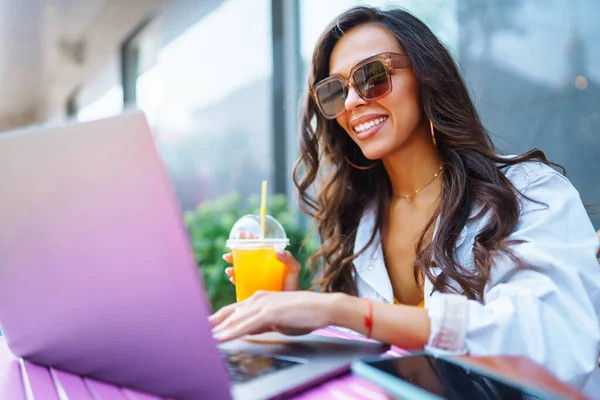 stock image Young and cheerful woman on the terrace chatting on Skype, talking on a webcam, having a video conference.Female is sitting in a modern cafe and working on a laptop. Freelancer, education concept.