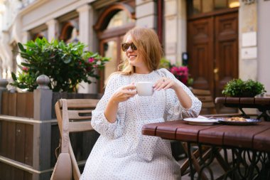 Sıcak bir kafenin verandasında kahve içen gülümseyen kadın. Turist kadın, bir fincan kahveyle şehir sokaklarının mimarisini ve sulama havasını seviyor. Turizm, seyahat ve tatil kavramı.