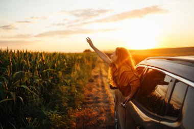 Genç mutlu kadın arabanın camından sarkıyor günbatımının tadını çıkarıyor. Aktif yaşam tarzı, seyahat, turizm, doğa.