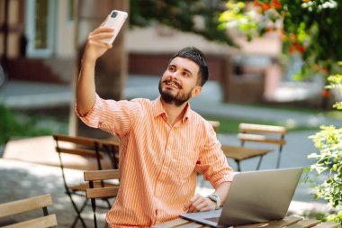 Sokakta bir kafede çalışan, dizüstü bilgisayarı ve telefonu olan gülümseyen genç bir adam. Serbest iş konsepti. İş, blog yazmak, serbest çalışmak, eğitim kavramı. Modern yaşam tarzı.