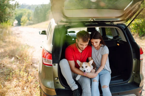 Lovely young couple with a dog sits in the back seat of a car. Happy man with a woman and his beloved dog travel together. Concept of active lifestyle, travel, weekend, relax.