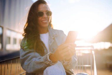 Akıllı telefonlu gülümseyen kadın, mavi duvara karşı alışveriş arabasıyla güneşli bir günün tadını çıkarıyor. Afrikalı görünümlü mutlu kadın alışverişe gidiyor. Yaşam tarzı, tüketim, satış kavramı.