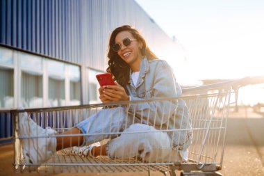 Akıllı telefonlu gülümseyen kadın, mavi duvara karşı alışveriş arabasıyla güneşli bir günün tadını çıkarıyor. Afrikalı görünümlü mutlu kadın alışverişe gidiyor. Yaşam tarzı, tüketim, satış kavramı.