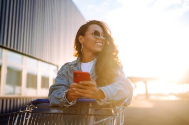 Akıllı telefonlu gülümseyen kadın, mavi duvara karşı alışveriş arabasıyla güneşli bir günün tadını çıkarıyor. Afrikalı görünümlü mutlu kadın alışverişe gidiyor. Yaşam tarzı, tüketim, satış kavramı.