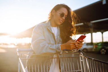 Akıllı telefonlu gülümseyen kadın, mavi duvara karşı alışveriş arabasıyla güneşli bir günün tadını çıkarıyor. Afrikalı görünümlü mutlu kadın alışverişe gidiyor. Yaşam tarzı, tüketim, satış kavramı.