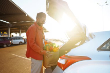 Otoparkta elinde kağıt torba Macar ürünleri olan bir adam. Arabanın yanında çantası olan genç bir adam. Zorlu yaşam tarzı kavramı, tüketim, satış, alışveriş.