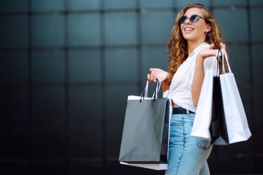 Alışveriş çantalı mutlu bir kadın sokakta güneşli havanın tadını çıkarıyor. Alışveriş merkezinden alışveriş yapan genç bir kadın. Yaz alışverişi. Yaşam tarzı, alışveriş, satış..
