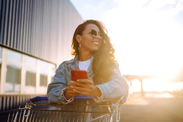 Uśmiechnięta Kobieta Smartfonem Wózkiem Zakupy Przy Niebieskiej Ścianie Bawi Się — Zdjęcie stockowe