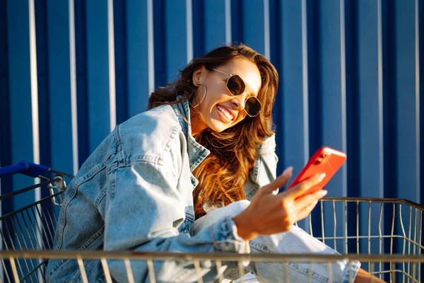 Smiling woman with a smartphone, with a shopping trolley against a blue wall has fun, enjoying sunny day. Happy woman of African appearance goes shopping. Concept of lifestyle, consumerism, sale.