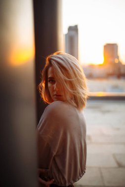 Şık bir kadın gün batımında çatıdan şehrin inanılmaz manzarasını izler. Çatıda poz veren genç bir kadın. Tarz, moda veya özgürlük konsepti, yaşam tarzı..