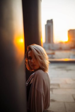 Şık bir kadın gün batımında çatıdan şehrin inanılmaz manzarasını izler. Çatıda poz veren genç bir kadın. Tarz, moda veya özgürlük konsepti, yaşam tarzı..