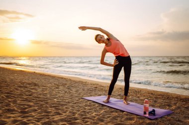 Gündoğumunda plajda yoga yapan modern genç bir kadın. Spor kadını dışarıda esneme hareketleri yapıyor. Aktif yaşam tarzı, spor eğitimi, yoga veya fitness kavramı.