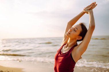 Gündoğumunda plajda yoga yapan modern genç bir kadın. Spor kadını dışarıda esneme hareketleri yapıyor. Aktif yaşam tarzı, spor eğitimi, yoga veya fitness kavramı.
