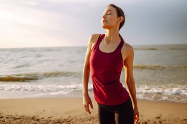 Gündoğumunda plajda yoga yapan modern genç bir kadın. Spor kadını dışarıda esneme hareketleri yapıyor. Aktif yaşam tarzı, spor eğitimi, yoga veya fitness kavramı.