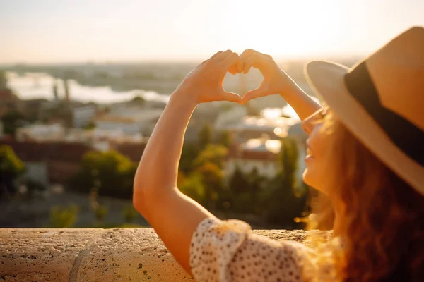 Woman tourist holding hands in the shape of heart looking at the panoramic landscape of the city. A young woman enjoys the freedom and beauty of the city at dawn. Active lifestyle concept and rest.