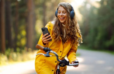 Kulaklığı ve akıllı telefonu olan mutlu genç bir kadın güneşli bir parkta bisiklet sürüyor müzik dinliyor, podcast yapıyor. Rahatla, doğa konsepti. Yaşam biçimi.