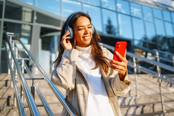 Optymistyczna Kobieta Nosząca Słuchawki Trzymająca Telefon Zewnątrz Młoda Kobieta Słucha — Zdjęcie stockowe