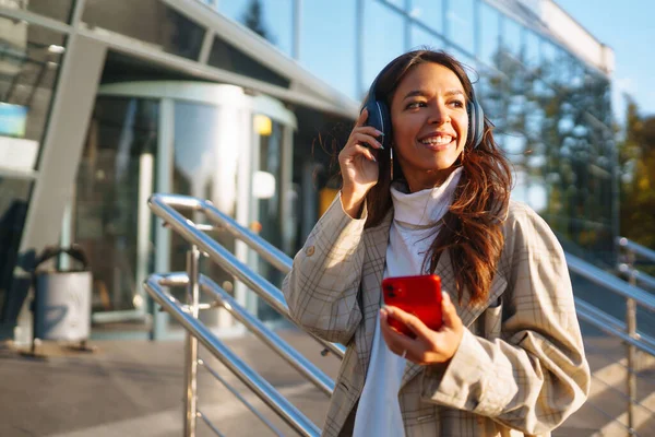 Optymistyczna Kobieta Nosząca Słuchawki Trzymająca Telefon Zewnątrz Młoda Kobieta Słucha — Zdjęcie stockowe
