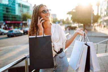Güneşli bir sokakta telefonla alışveriş yapan şık genç bir kadın. Güzel bir kadın cep telefonu kullanıyor ve alışveriş torbaları tutuyor. Alışveriş, satış, tüketim kavramı. Etkin yaşam biçimi.