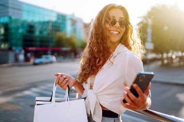 Güneşli bir sokakta telefonla alışveriş yapan şık genç bir kadın. Güzel bir kadın cep telefonu kullanıyor ve alışveriş torbaları tutuyor. Alışveriş, satış, tüketim kavramı. Etkin yaşam biçimi.