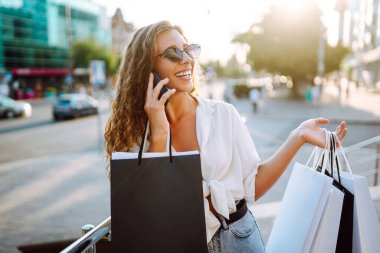 Güneşli bir sokakta telefonla alışveriş yapan şık genç bir kadın. Güzel bir kadın cep telefonu kullanıyor ve alışveriş torbaları tutuyor. Alışveriş, satış, tüketim kavramı. Etkin yaşam biçimi.