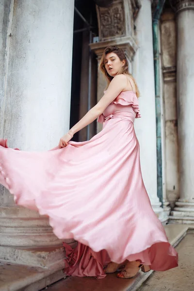 Fashion portrait of stylish young woman with wavy blond hair in an elegant pink dress. Model poses in a beautiful dress against the backdrop of classical architecture. Fashion and style concept.