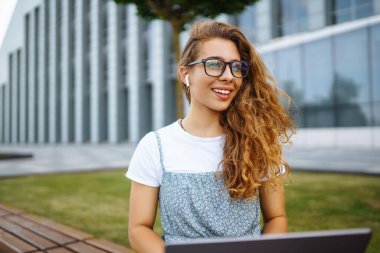 Gözlüklü, kablosuz kulaklıklı ve dizüstü bilgisayarı olan gülümseyen genç bir kadın bir şehir binasının arka planında çalışıyor ya da çalışıyor. Eğitim, iş, blog veya serbest çalışma kavramı.