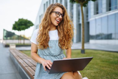 Gözlüklü, kablosuz kulaklıklı ve dizüstü bilgisayarı olan gülümseyen genç bir kadın bir şehir binasının arka planında çalışıyor ya da çalışıyor. Eğitim, iş, blog veya serbest çalışma kavramı.
