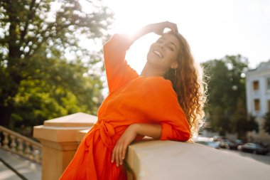 Parlak turuncu elbiseyle şehir manzarasının arka planında poz veren güzel genç bir kadın. Parlak elbiseli kadın turist güneşli bir günde şehri geziyor. Yaşam tarzı, moda, seyahat kavramı.