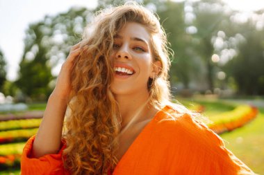 Parlak turuncu elbiseyle şehir manzarasının arka planında poz veren güzel genç bir kadın. Parlak elbiseli kadın turist güneşli bir günde şehri geziyor. Yaşam tarzı, moda, seyahat kavramı.