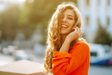 Parlak turuncu elbiseyle şehir manzarasının arka planında poz veren güzel genç bir kadın. Parlak elbiseli kadın turist güneşli bir günde şehri geziyor. Yaşam tarzı, moda, seyahat kavramı.