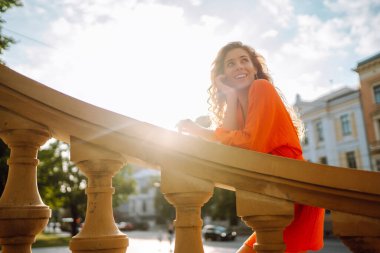 Parlak turuncu elbiseyle şehir manzarasının arka planında poz veren güzel genç bir kadın. Parlak elbiseli kadın turist güneşli bir günde şehri geziyor. Yaşam tarzı, moda, seyahat kavramı.