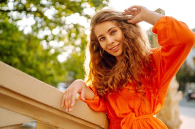 Parlak turuncu elbiseyle şehir manzarasının arka planında poz veren güzel genç bir kadın. Parlak elbiseli kadın turist güneşli bir günde şehri geziyor. Yaşam tarzı, moda, seyahat kavramı.