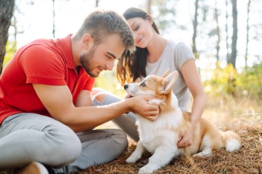 Young couple together with their beloved pet on a walk on a sunny day. Happy man and woman together with their corgi dog have together. The concept of an active lifestyle, travel, love and vacation. clipart