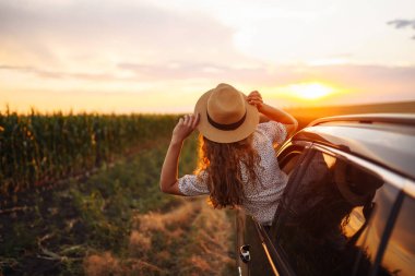 Şapkalı genç kadın, pencereden dışarı sarkan araba yolculuğundan hoşlanıyor. Yazın, güzel bir turist kadın arabayla seyahat eder ve altın gün batımının tadını çıkarır. Aktif yaşam tarzı, seyahat, turizm kavramı.