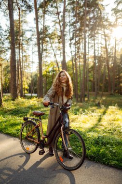 Güneşli bir parkta bisiklet süren şık bir genç kadın. Paltoyla bukleli güzel bir turist bisiklet sürüyor ve bahar havasının tadını çıkarıyor. Doğa kavramı, rahatla, aktif yaşam tarzı.