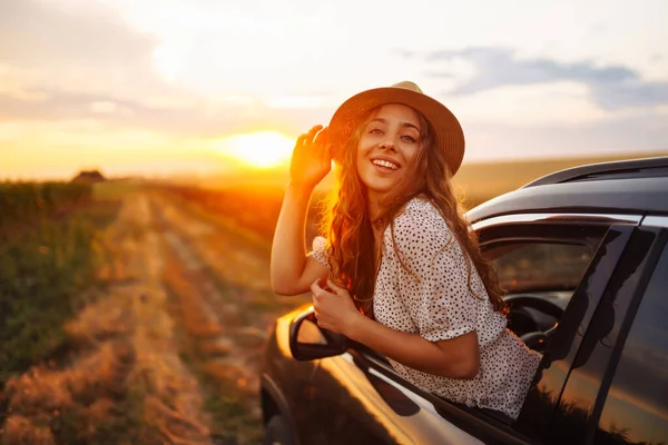 戴帽子的年轻而快乐的女人喜欢开车 靠在窗外 一位漂亮的旅游妇女开车旅行 欣赏金色的落日 积极生活方式 旅游的概念 — 图库照片