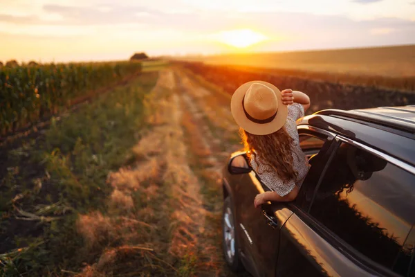 戴帽子的年轻而快乐的女人喜欢开车 靠在窗外 一位漂亮的旅游妇女开车旅行 欣赏金色的落日 积极生活方式 旅游的概念 — 图库照片