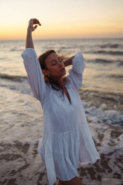 Deniz kıyısında beyaz bir elbise içinde poz veren güzel bir manken. Genç bir kadın kumsalda inanılmaz bir gün doğumunun tadını çıkarıyor. Yaşam tarzı. Tatil, seyahat kavramı. 