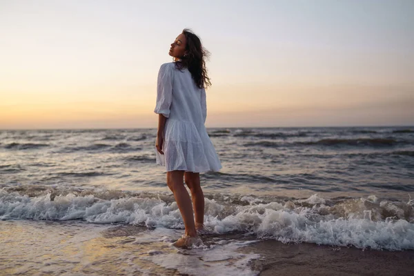 繊細な白いドレスに身を包んだ海岸沿いの美しいモデル 若い女性がビーチで信じられないほどの日の出を楽しんでいます ライフスタイル 旅行のコンセプト — ストック写真
