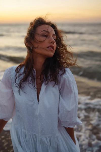 繊細な白いドレスに身を包んだ海岸沿いの美しいモデル 若い女性がビーチで信じられないほどの日の出を楽しんでいます ライフスタイル 旅行のコンセプト — ストック写真