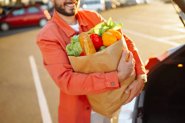 Stylish man with paper bags of vegetarian products in the parking lot near his car. Shopping for healthy vegetarian food in men\'s hand. Time for shopping.