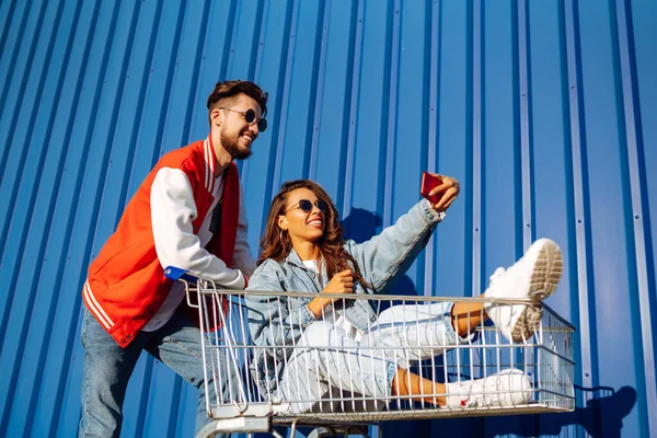 快乐的男人和女人在购物车上玩着 拿着手机 用智能手机拍着自拍 购物时间 年轻人的观念 自私的时间 — 图库照片