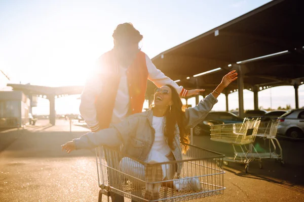幸せなカップルはカートに乗って楽しみを持っています 笑顔の男と女が買い物に行く ブラック フライデー ライフスタイルの概念 消費主義 — ストック写真