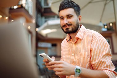 Bilgisayarda telefonla çalışırken gülümseyen mutlu erkek serbest yazar, online konferans. Genç adam sokak kafesinde çalışıyor. Serbest iş konsepti. Günlük, eğitim kavramı.