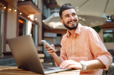 Bilgisayarda telefonla çalışırken gülümseyen mutlu erkek serbest yazar, online konferans. Genç adam sokak kafesinde çalışıyor. Serbest iş konsepti. Günlük, eğitim kavramı.