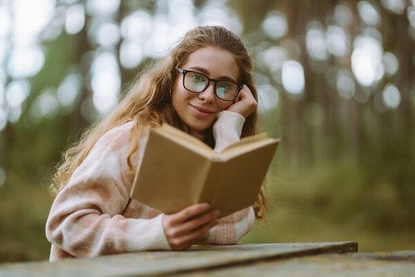 Happy young woman is sitting at table with book in beautiful forest. Beautiful woman is enjoying fresh air in forest reading book. Concept of relaxation, solitude with nature, lifestyle.